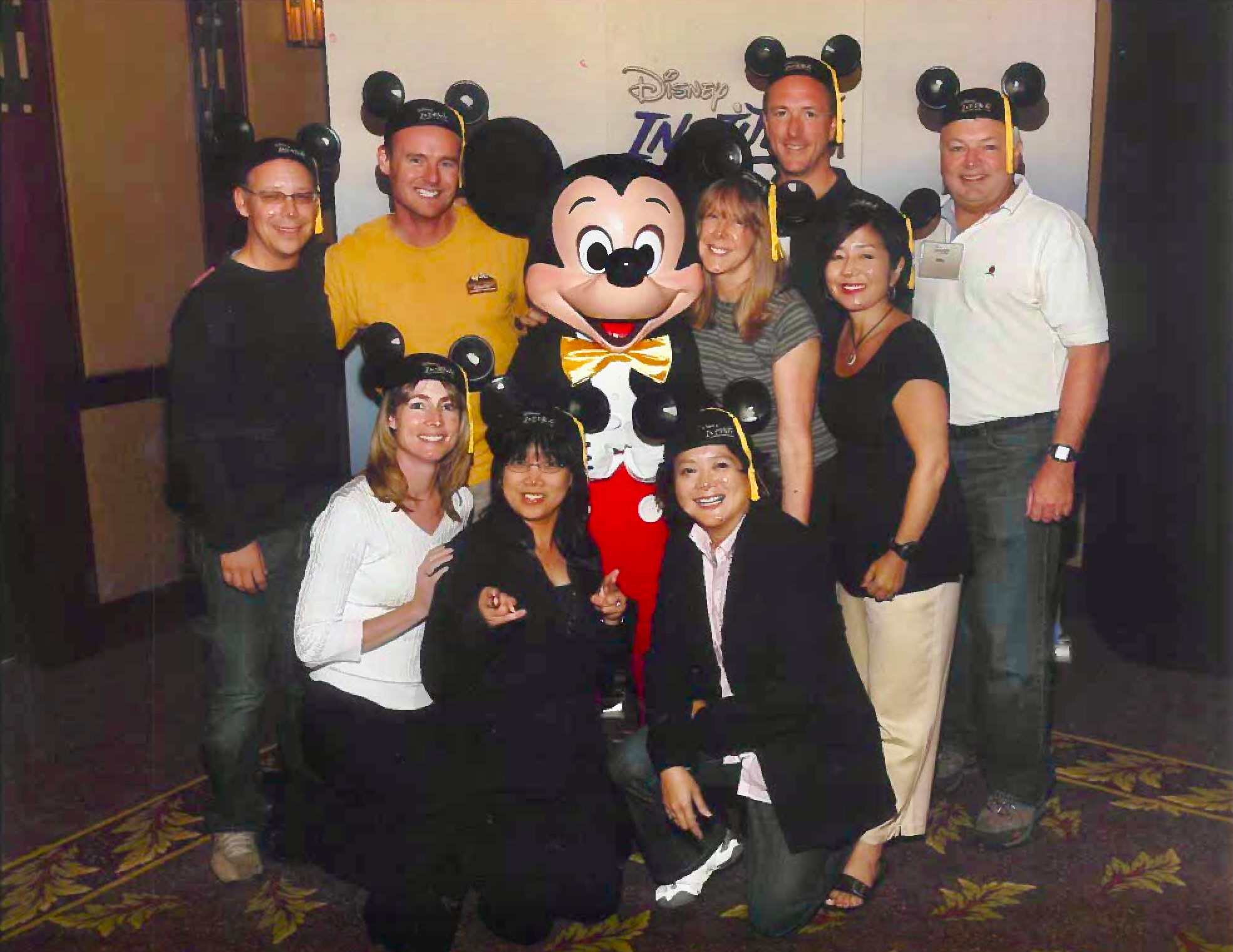 A group of employees huddle around Mickey Mouse while wearing Disney Mickey hats.