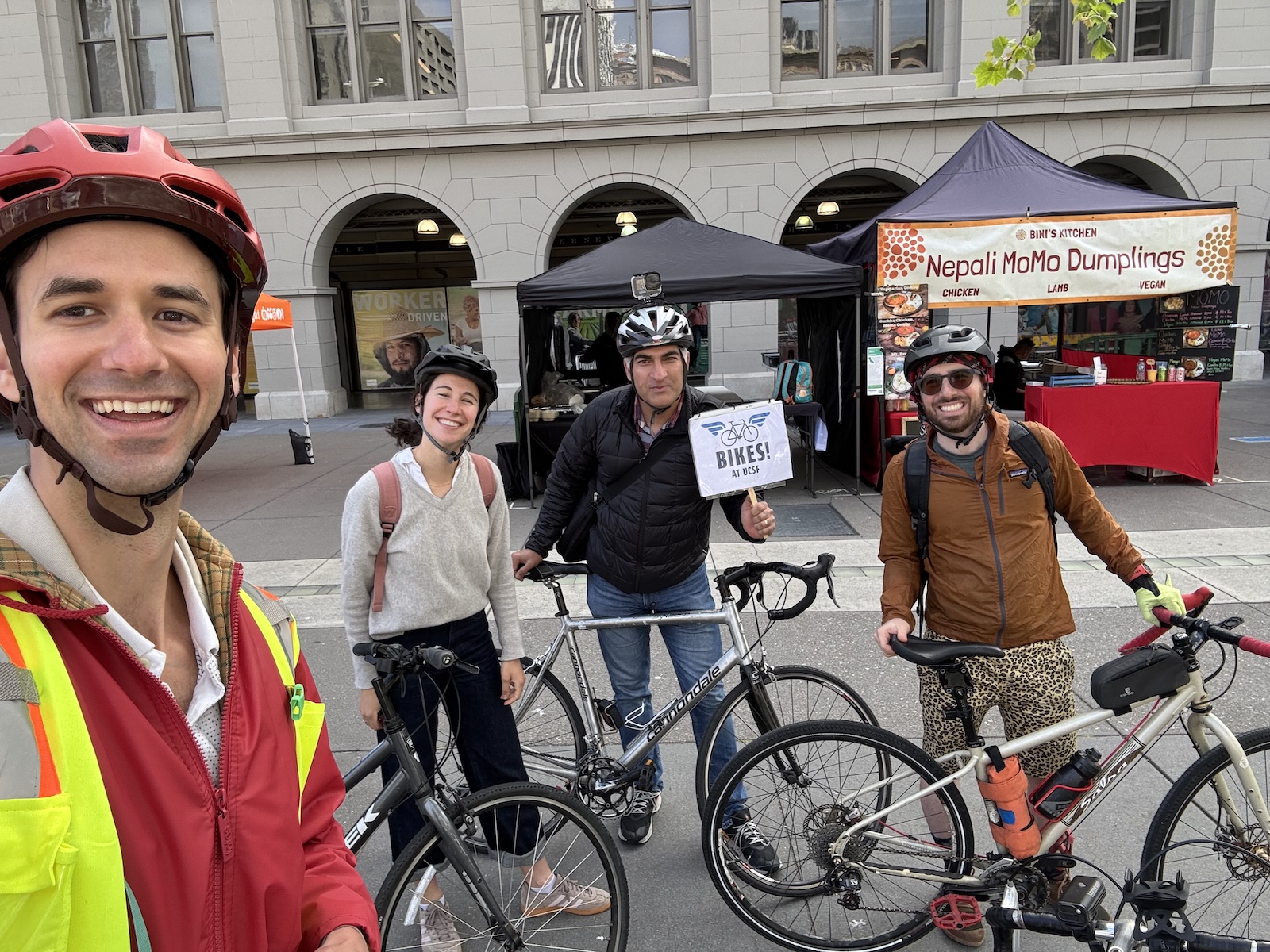 Cyclists gather in front of the Ferry Building, one holds a sign saying 'Bikes! at UCSF'