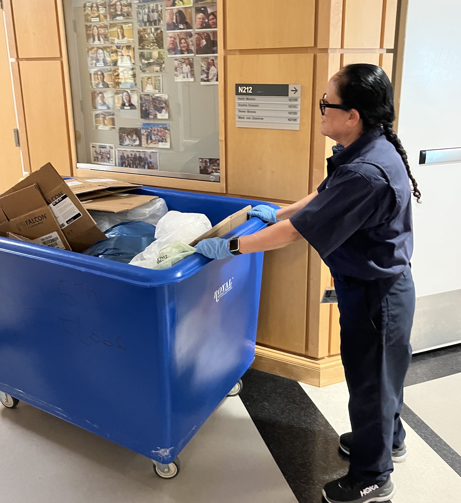 A woman in uniform pushes a blue recycling cart down a hallway.