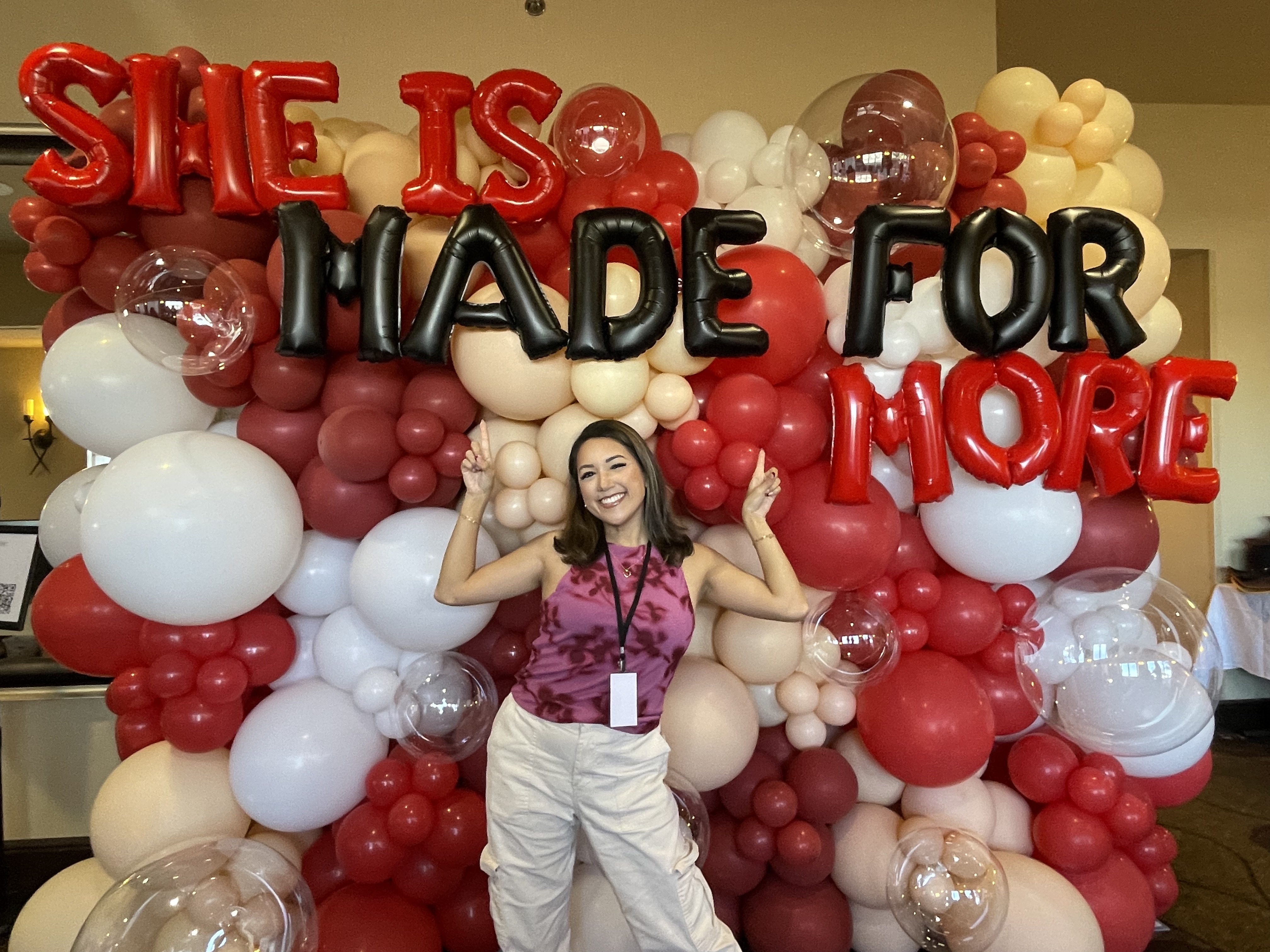 Aguilar poses in front of a read and white balloon display with the balloon letters spelling 'She is Made for More'