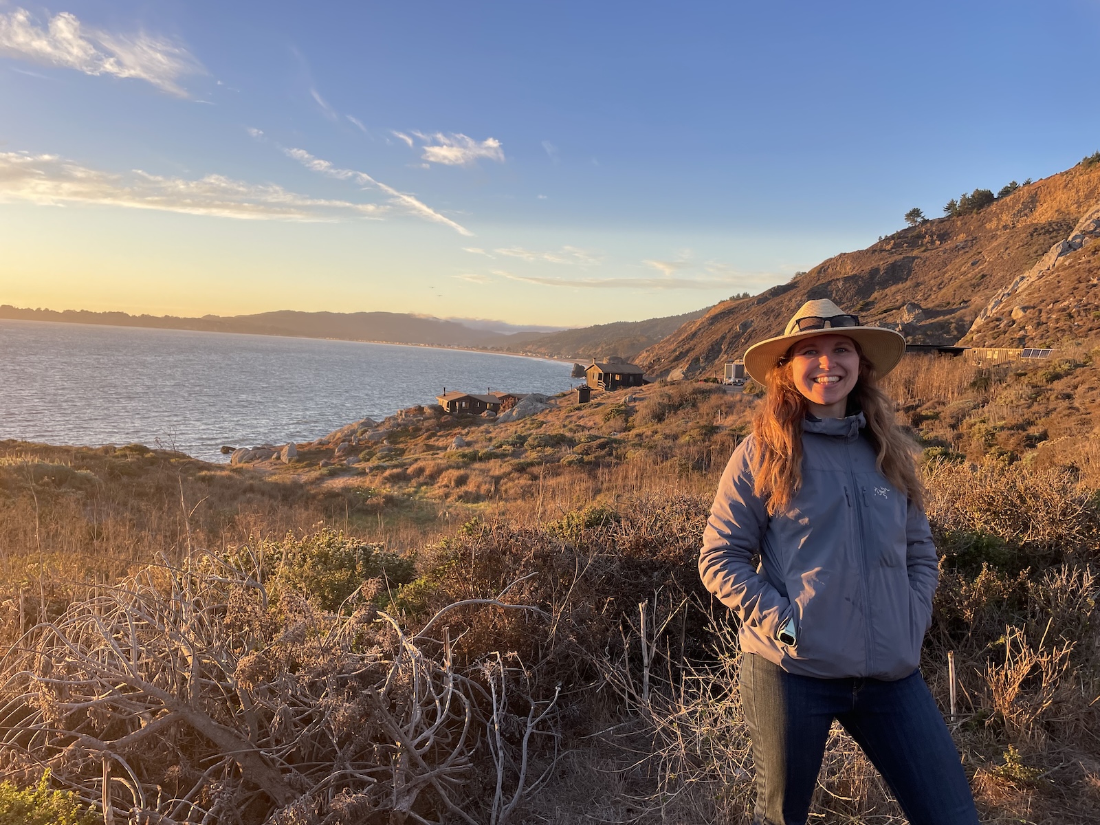 A woman stands on a trail wearing a hat and smiling