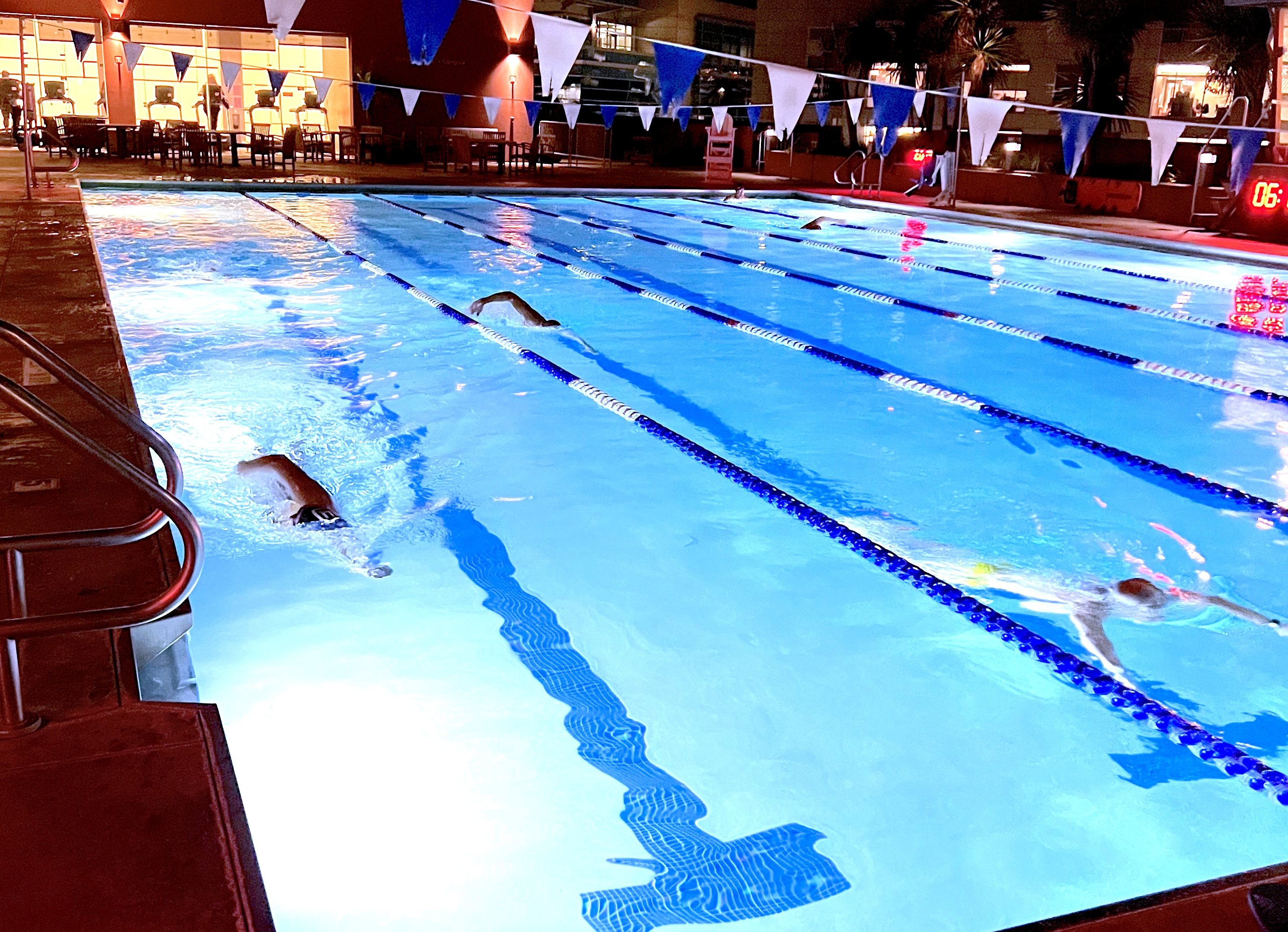 Three swimmers practice crawl stroke in the outdoor pool under blue and white flags.