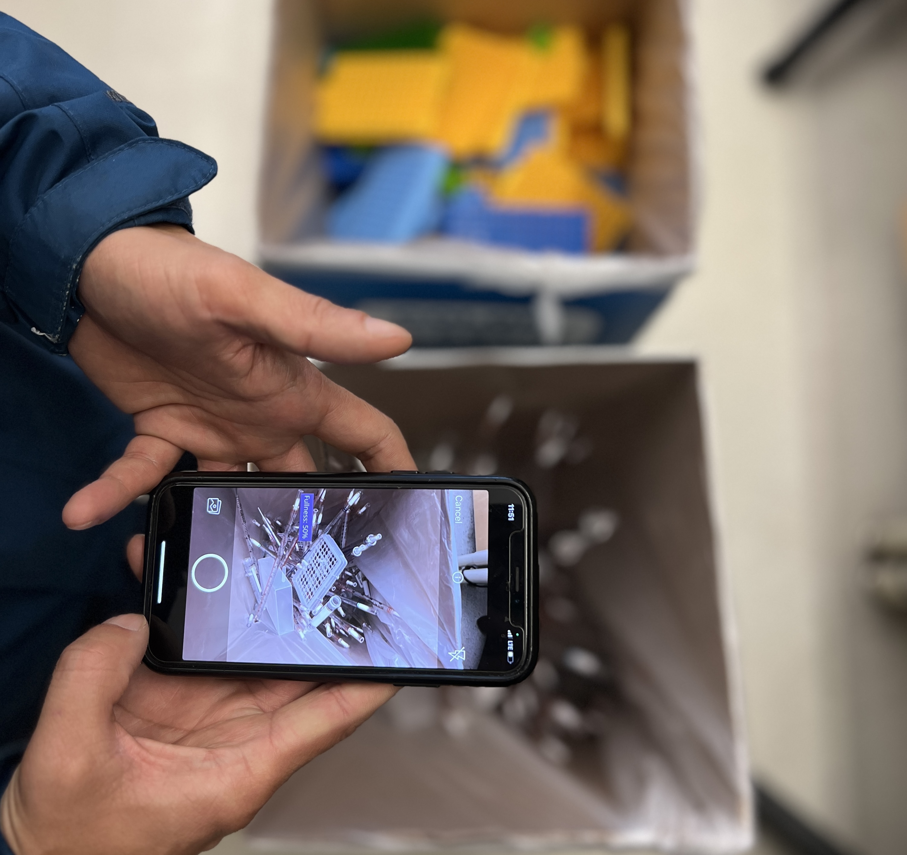 Facilities team member holds phone over recycling box to take a photo of lab syringes and plastics.
