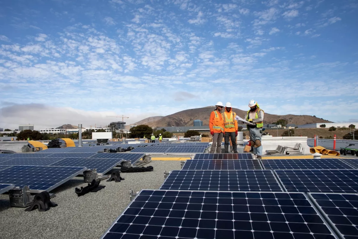 Workers in high-visibility safety gear and hard hats reviewing architectural or electrical blueprints on-site during installation of solar panels