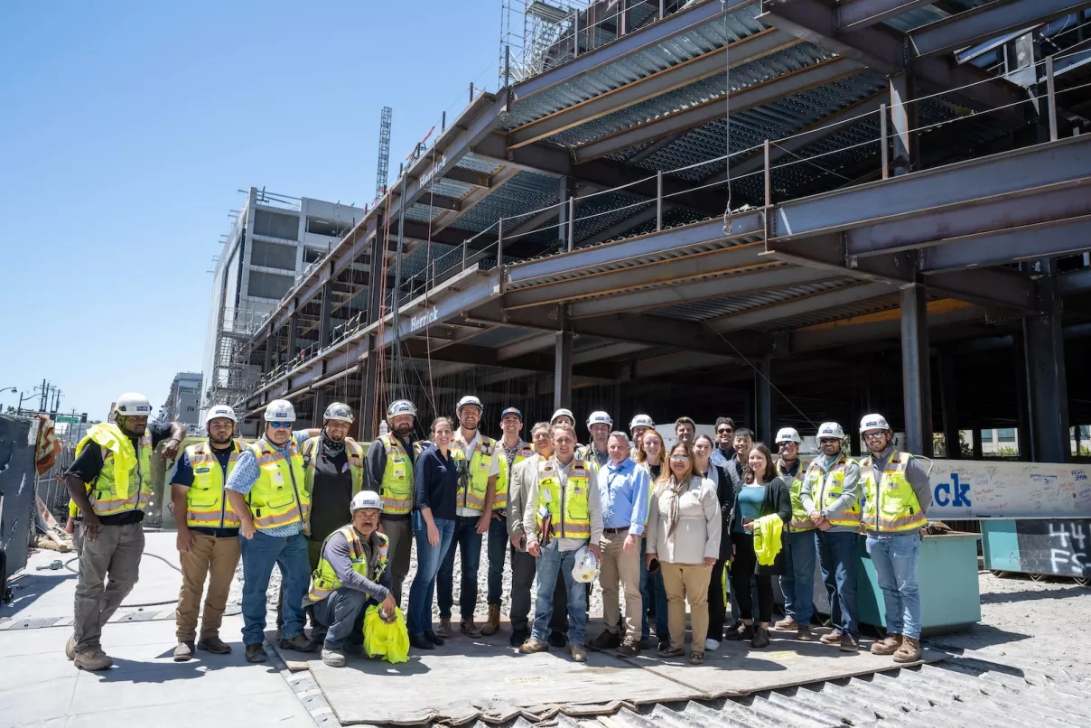 a group of construction workers and project team members posing in front of a multi-story steel-frame building structure on a sunny day